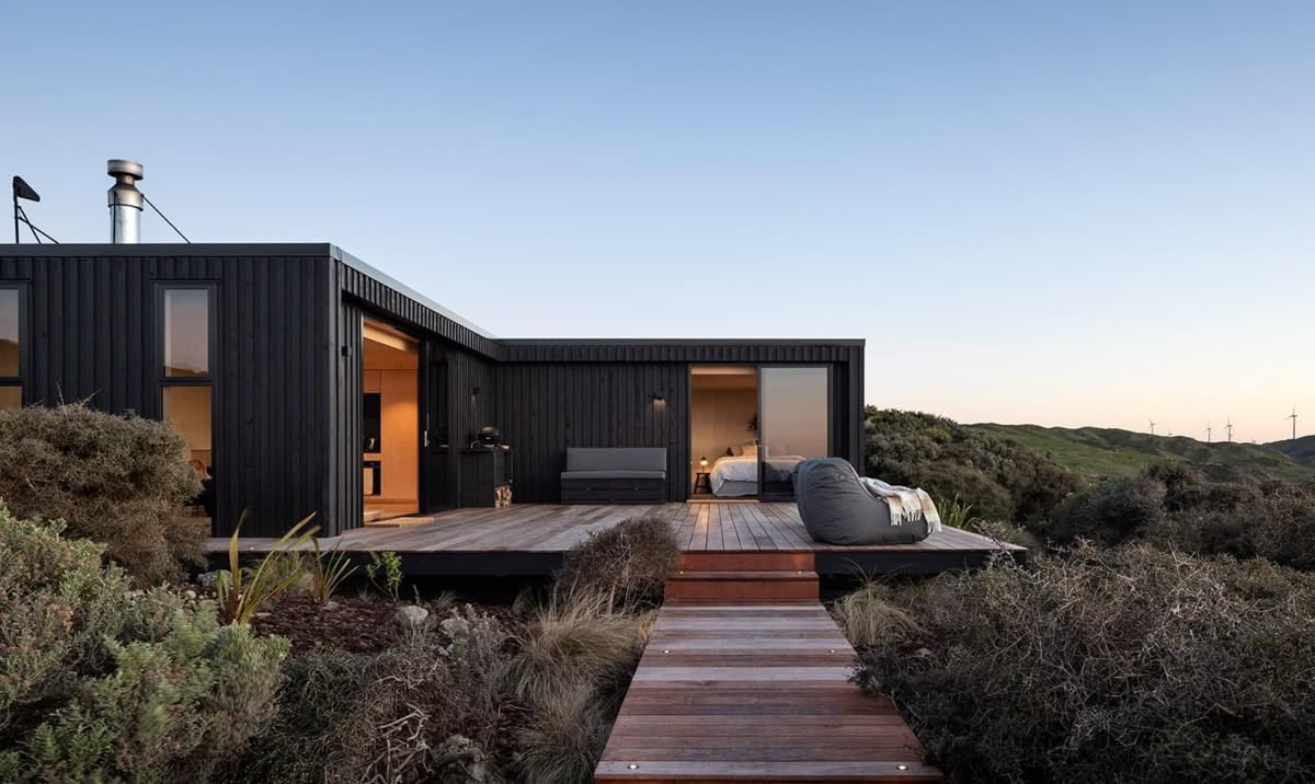 Vivienda modular de fachada oscura con deck de madera y grandes ventanales, integrada a un paisaje natural al atardecer