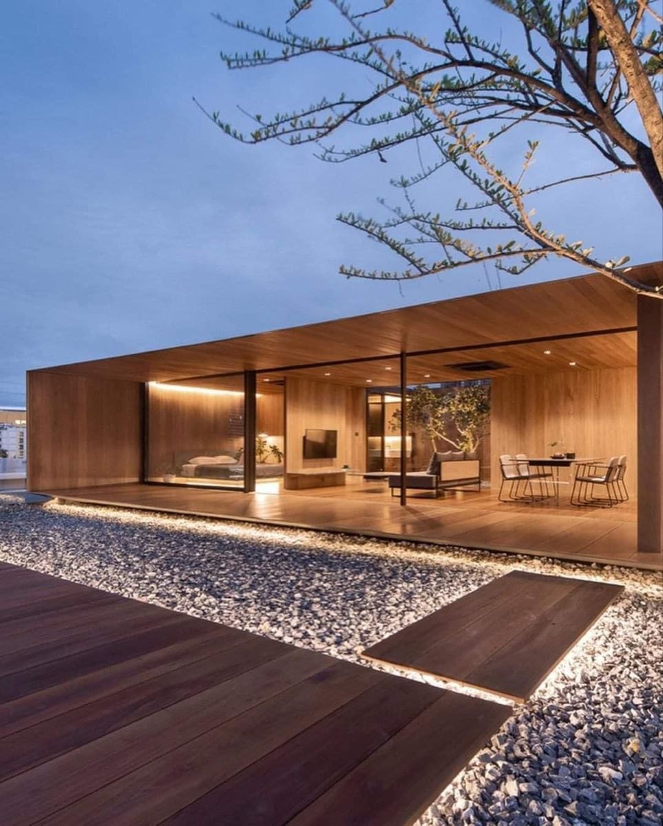 Pabellón de madera con grandes paños vidriados e iluminación cálida, rodeado de canto rodado y deck